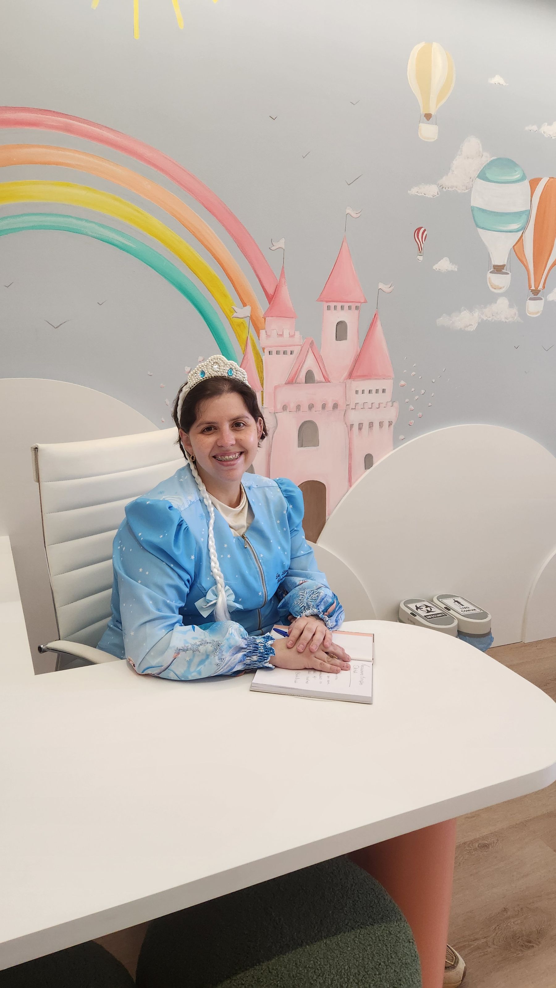 A Dra. Luiza Bretas, a Tia Lú, fantasiada de Elsa, sorrindo na mesa do consultório, com o mural do castelo e do arco-íris atrás